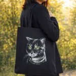 Tote-bag noir peint à la main avec portrait de chat tigré, porté par une femme élégante en promenade dans un bois ensoleillé.